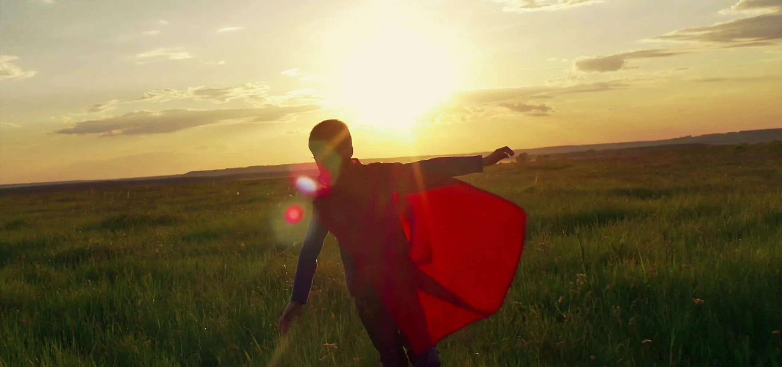 A child with a cape is playing in a meadow during the sunset